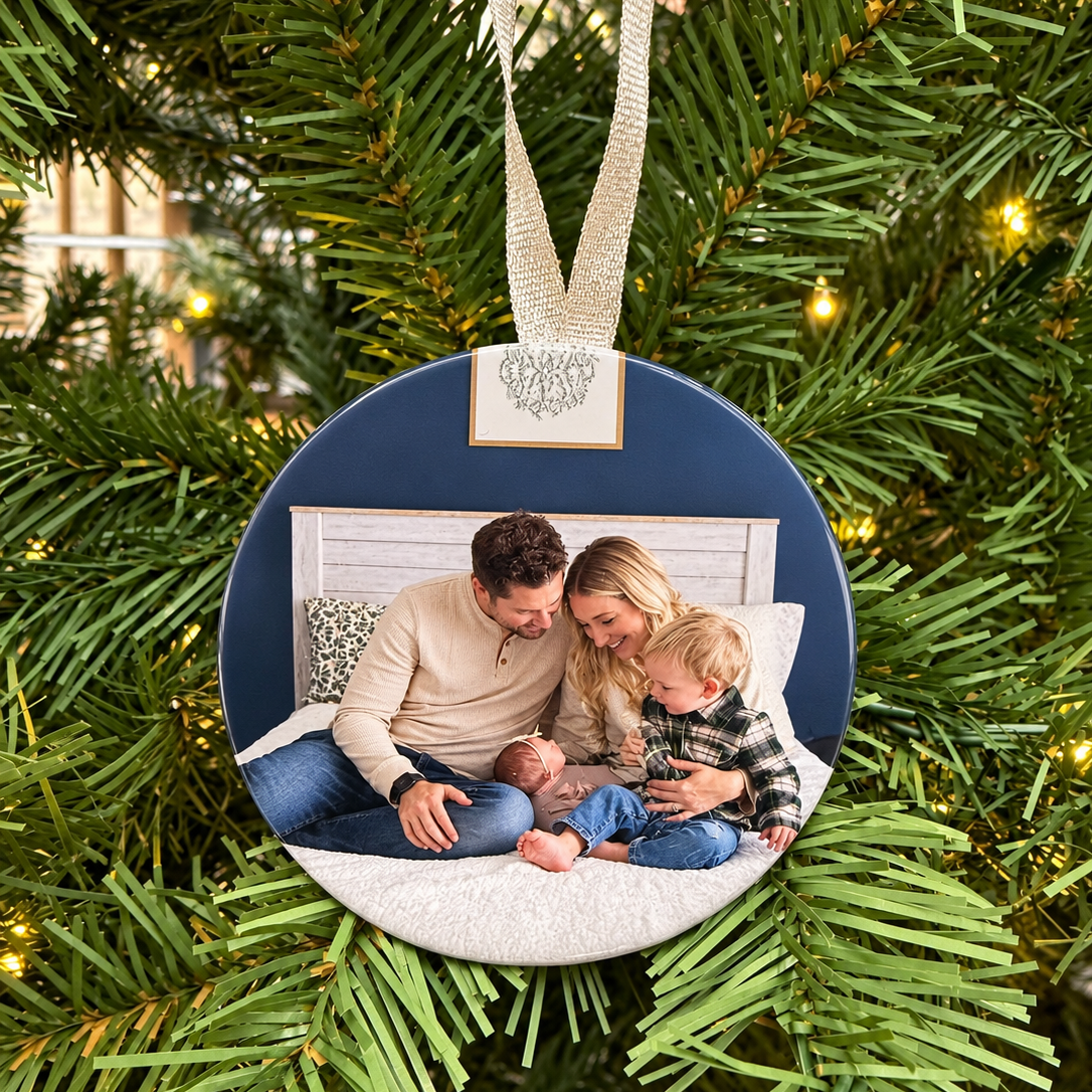 A round Christmas ornament hanging on a decorated Christmas tree. The ornament features a custom printed photo of a family of four sitting on a bench, with a navy blue background and a white textured floor.